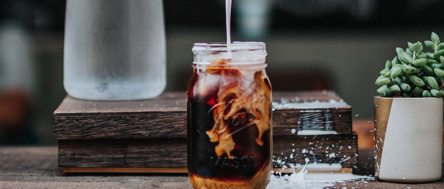 Cold brew coffee in a mason jar, milk being poured on top of it and a little bit of splashing